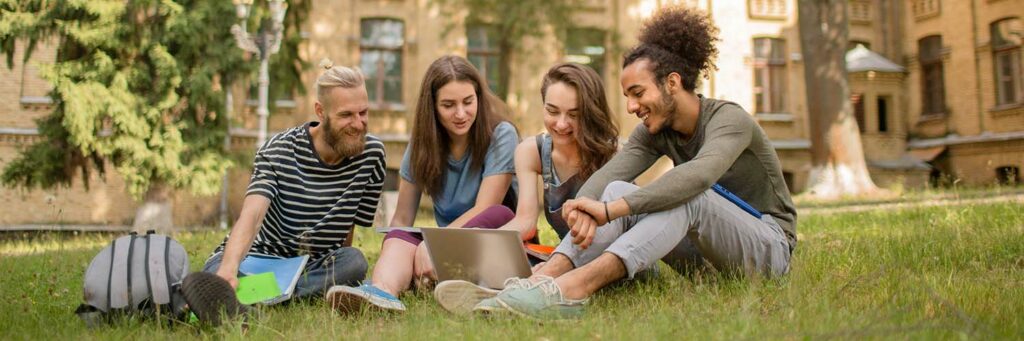 View on cheerful students sitting on grass on sunny day. Smiling college friends working with laptop on lawn.