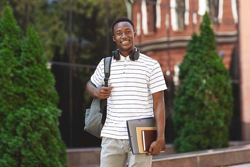 Happy smiling african american student guy with backpack and workbooks over university background, looking at camera and smiling