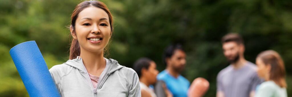fitness, sport and healthy lifestyle concept - happy smiling young asian woman with mat over group of people meeting for yoga class at summer park
