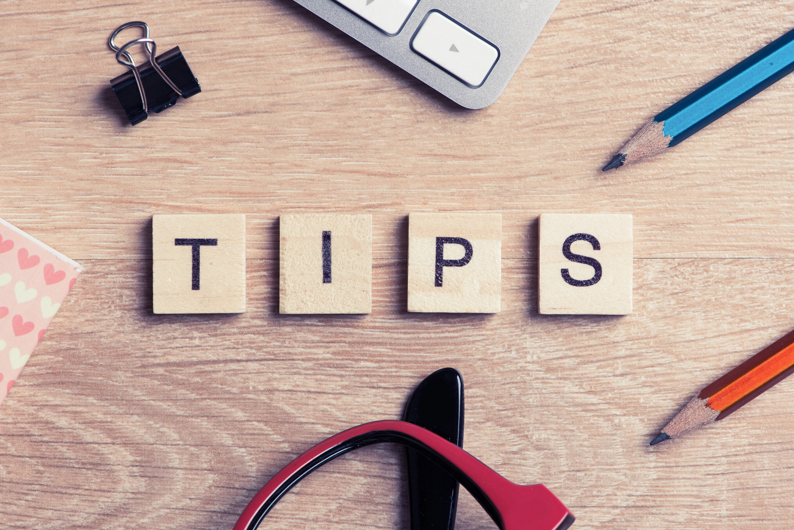 Corner of laptop with pencils, binder clip, glasses, and tiles that spell "TIPS" on desk.