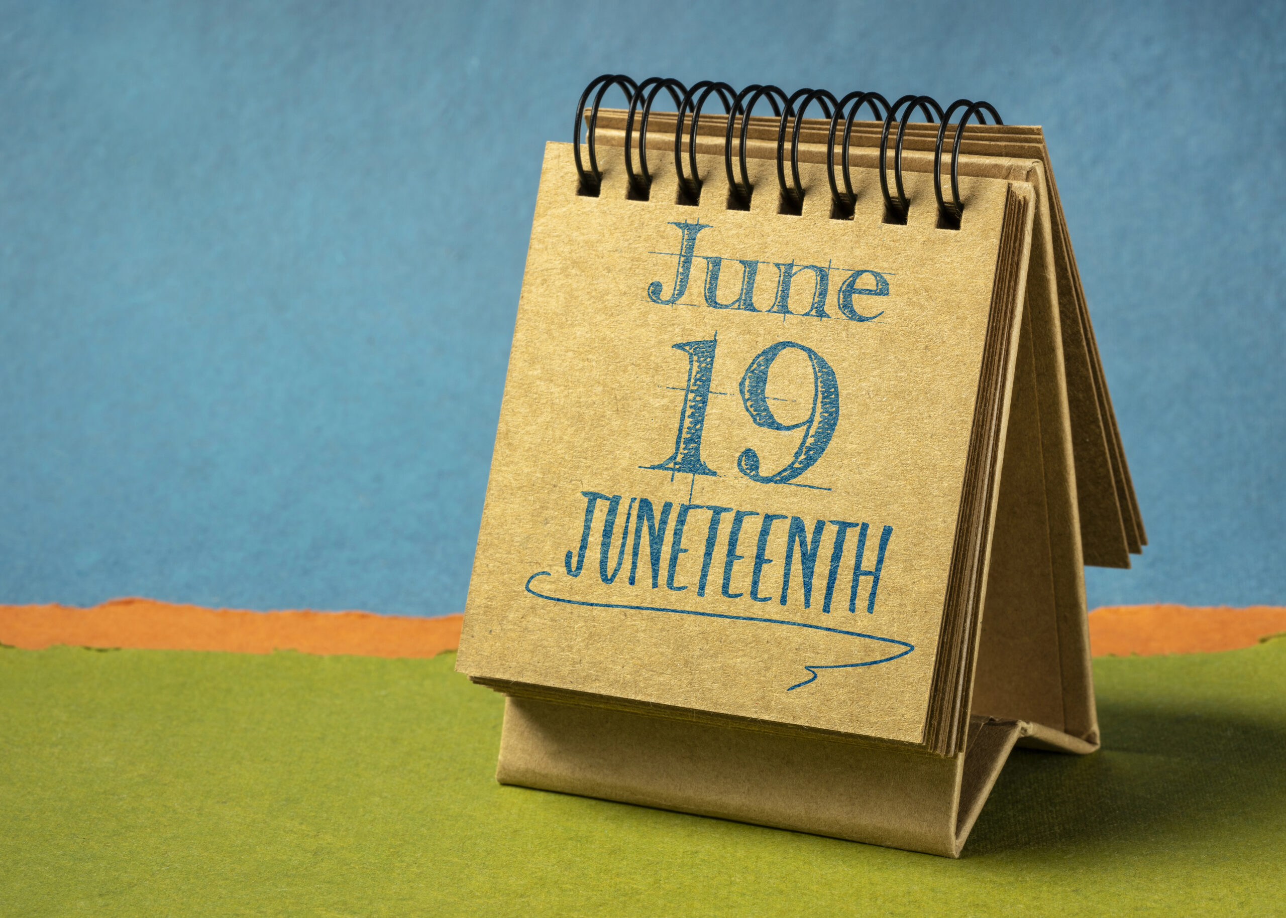 Stand-up desk calendar with June 19 Juneteenth on it against blue and green background.